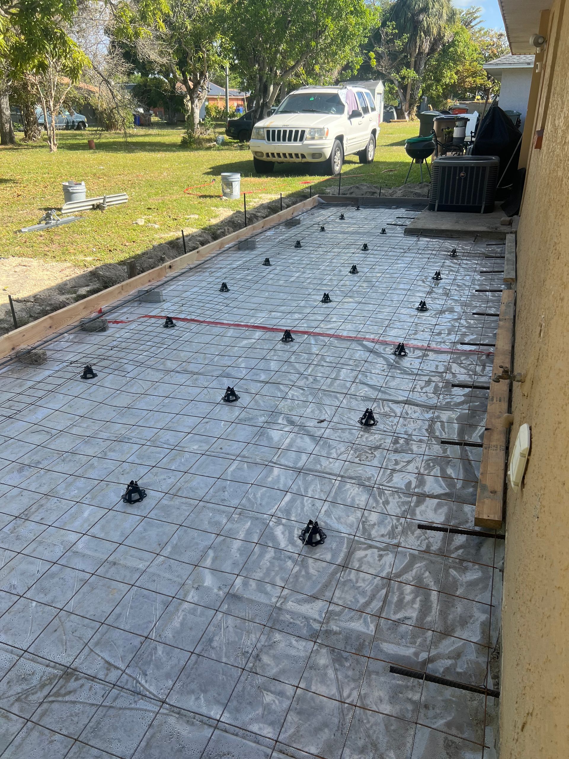Concrete slab construction: forms, rebar grid, plastic sheeting, and supports in a backyard setting with a vehicle.