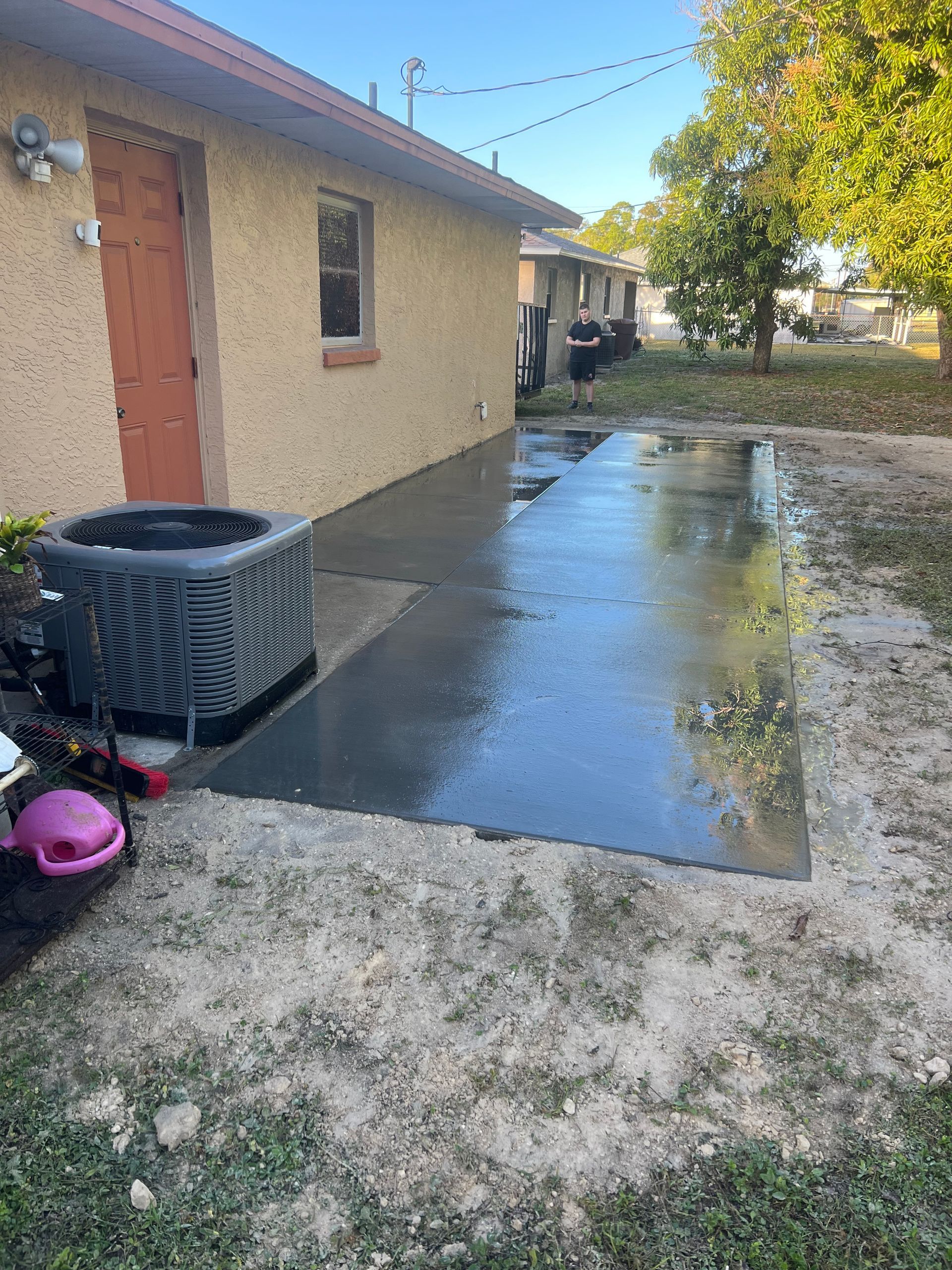 A wet concrete patio next to a beige building. An air conditioning unit and some yard items are visible.