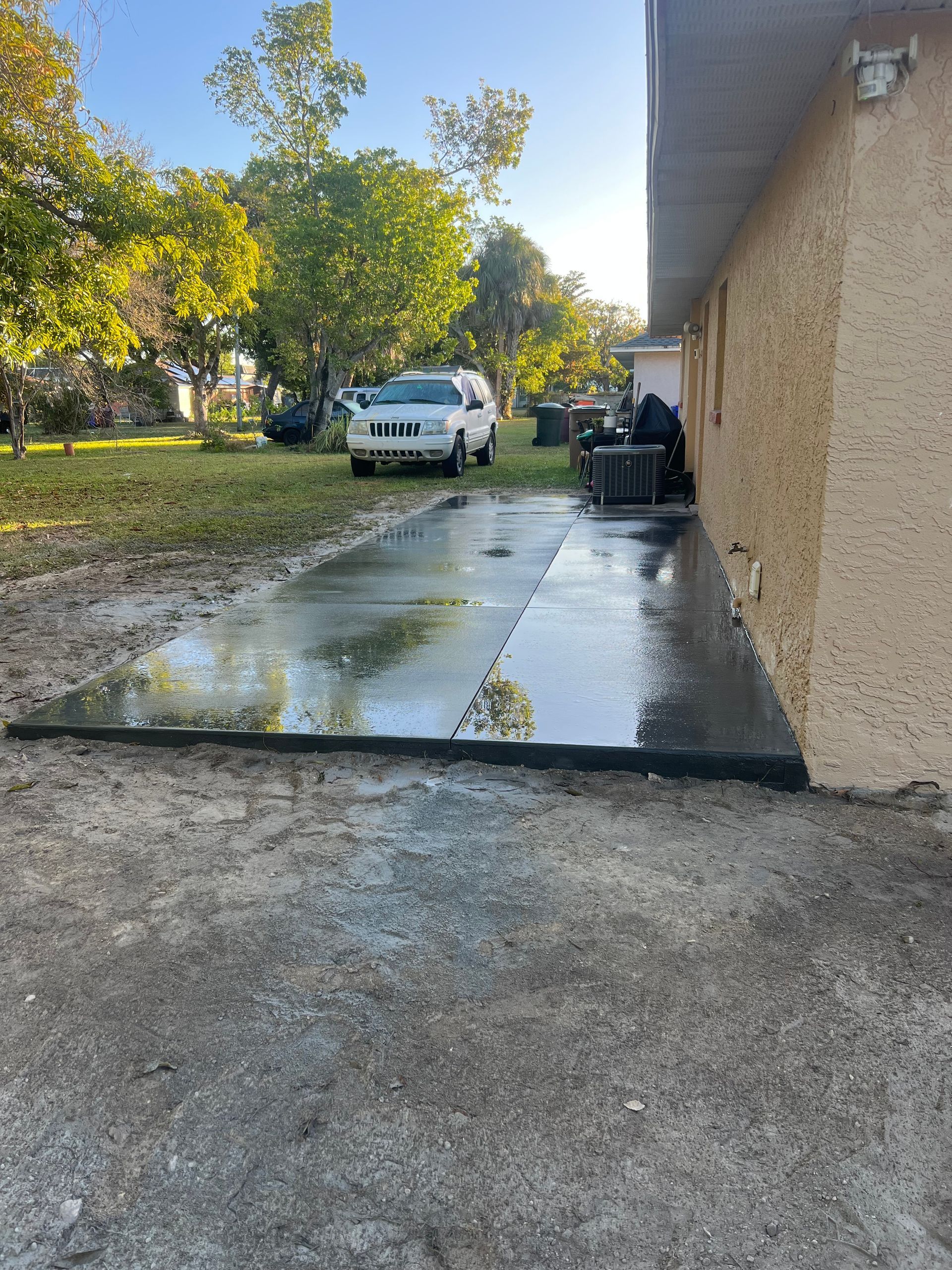 Wet concrete patio next to a beige building, with a car in the background and a grassy lawn.