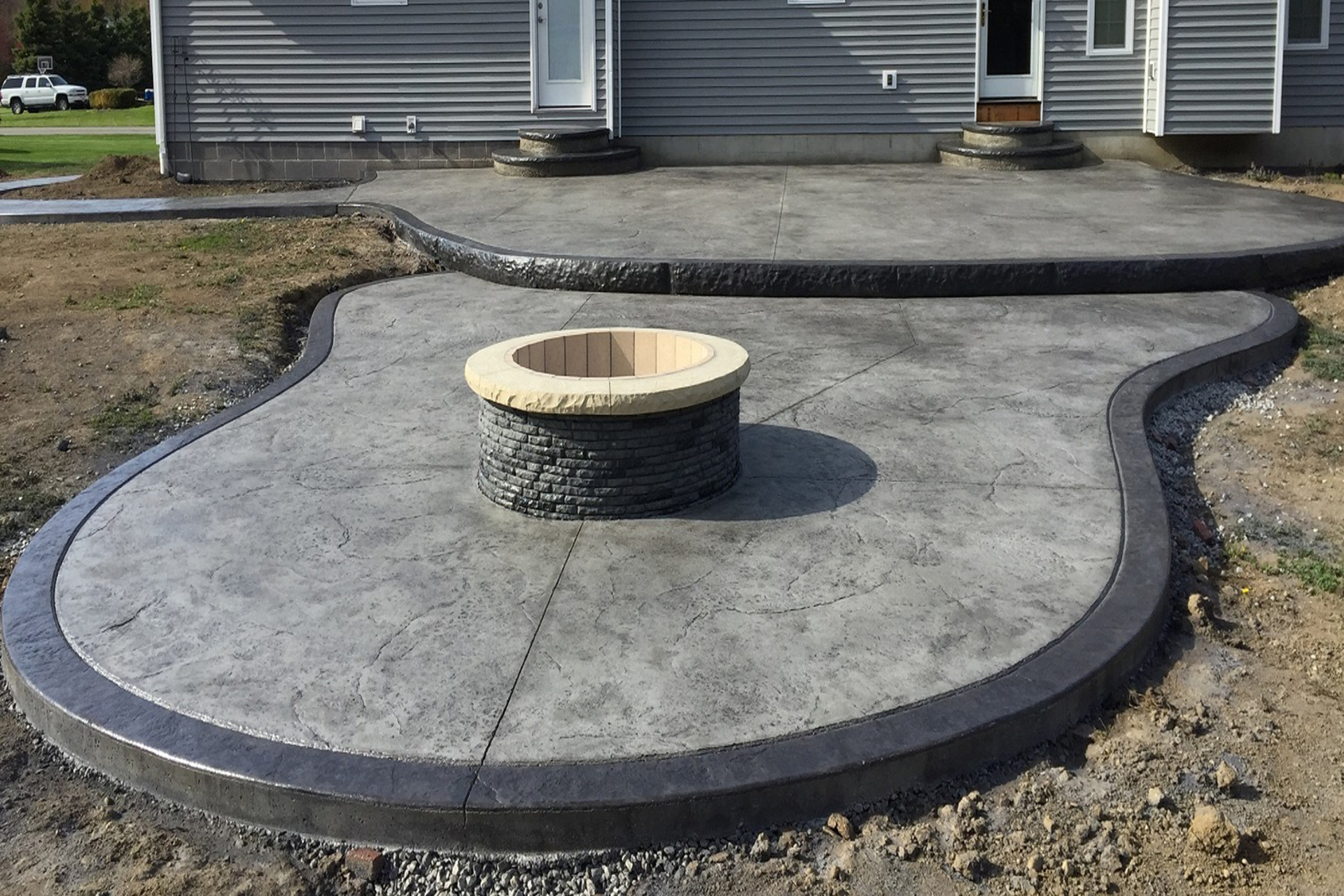 Concrete patio with fire pit, bordered with dark edging, in front of a gray house.