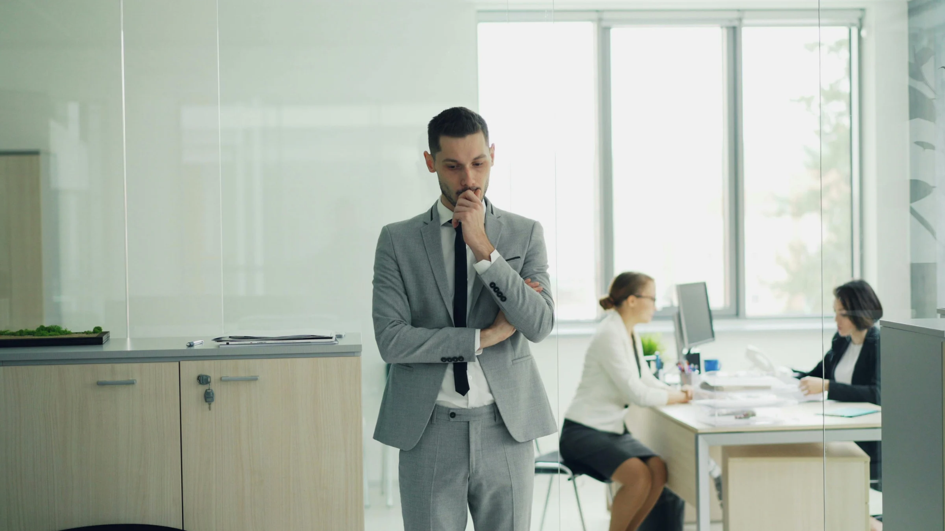 Businessman standing thoughtfully in a bright office, with two people working at a desk in the background