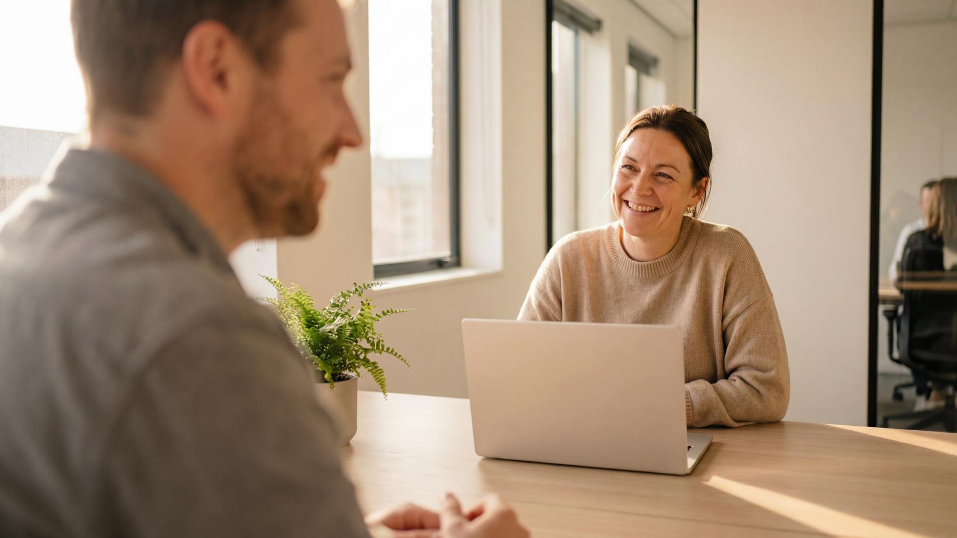 A smiling person sits at a desk with a laptop, engaged in a conversation with a colleague in a sunlit office.