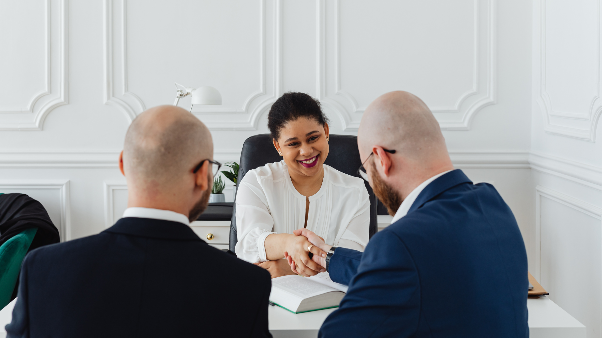 Woman shaking hands with a man at a desk, another man is on the left. White office, smiling faces.
