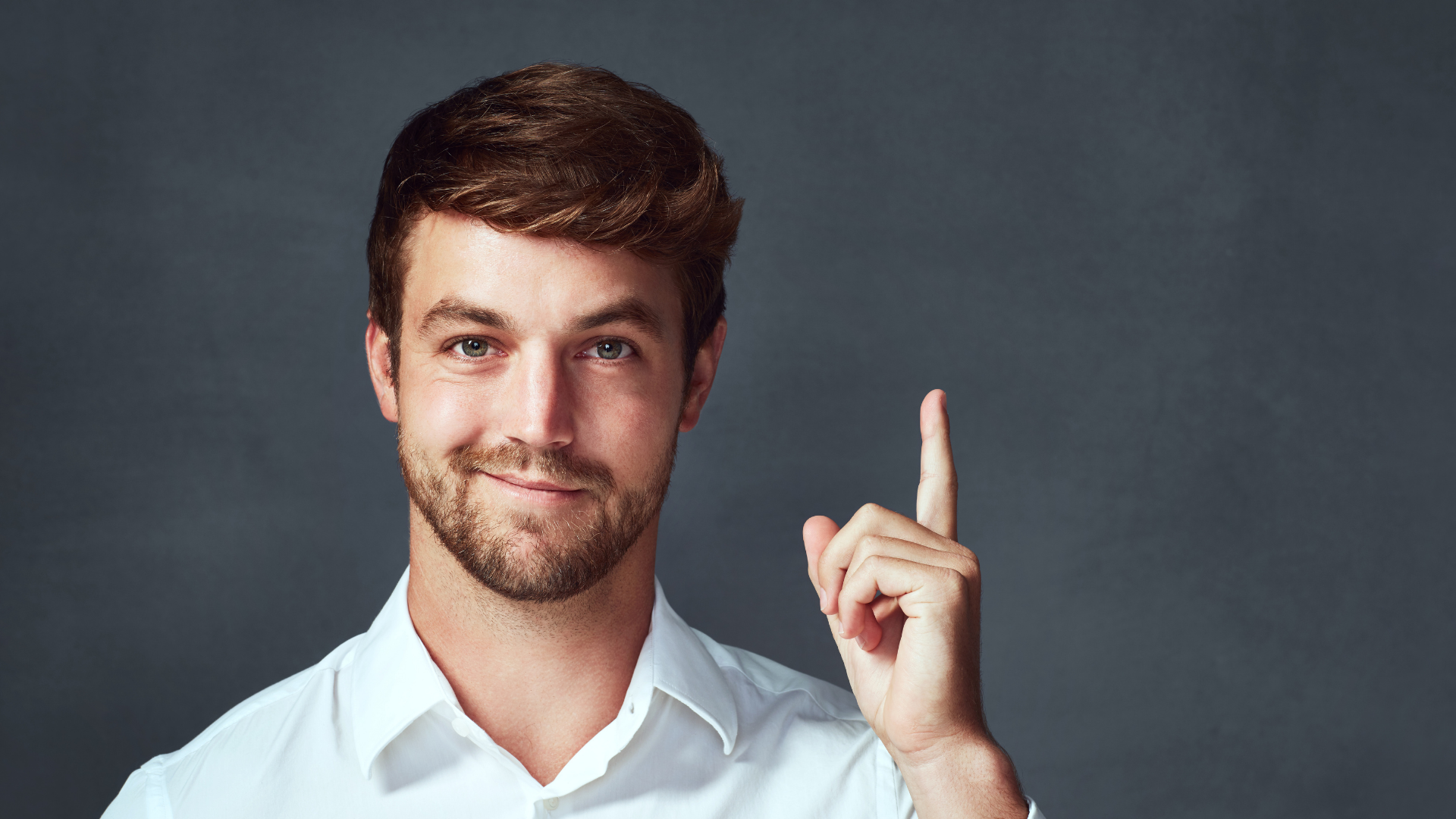 Man in white shirt smiling and pointing upward with one finger on a gray background