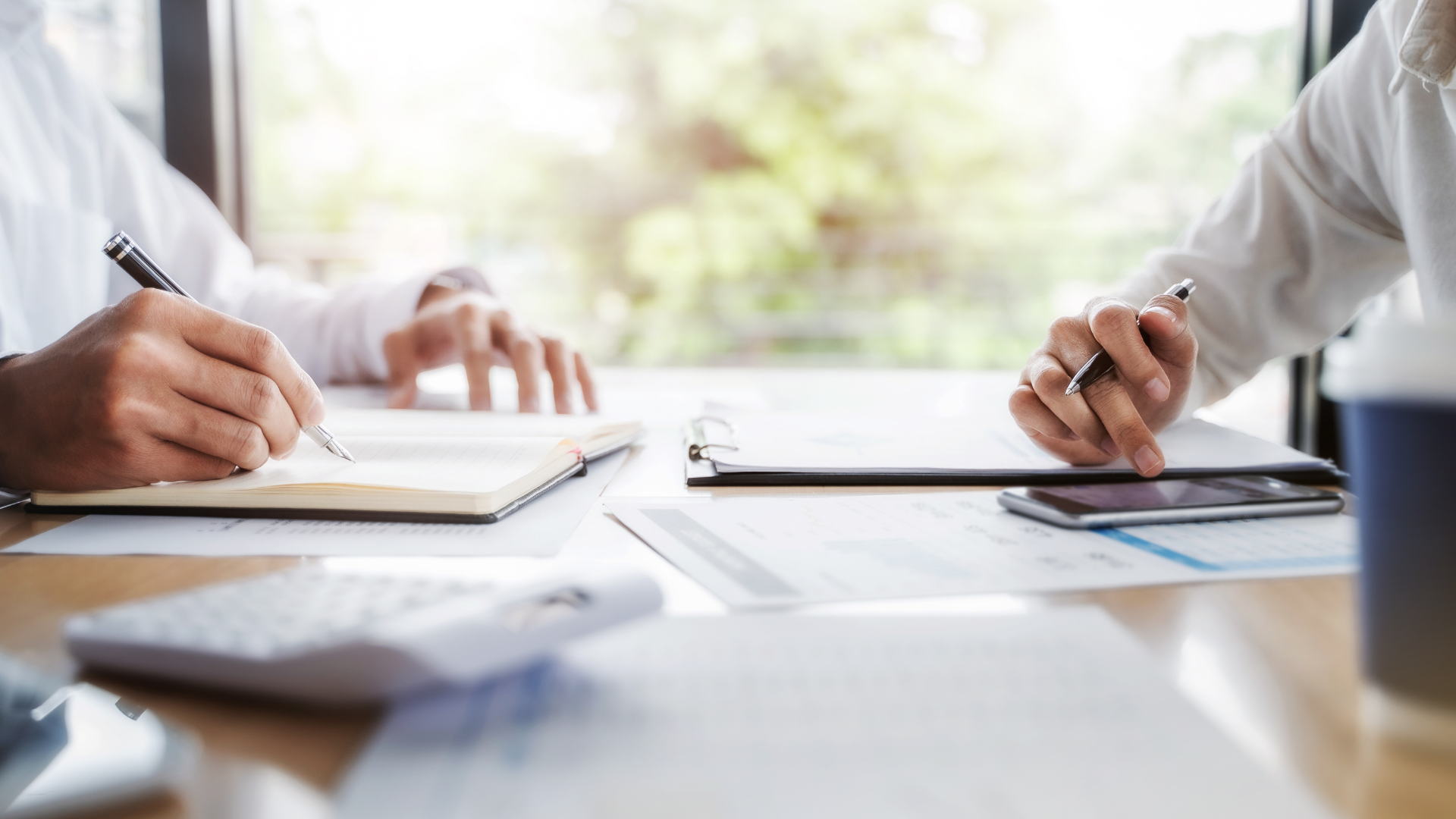 Two people writing at a bright office table with notebooks and a laptop