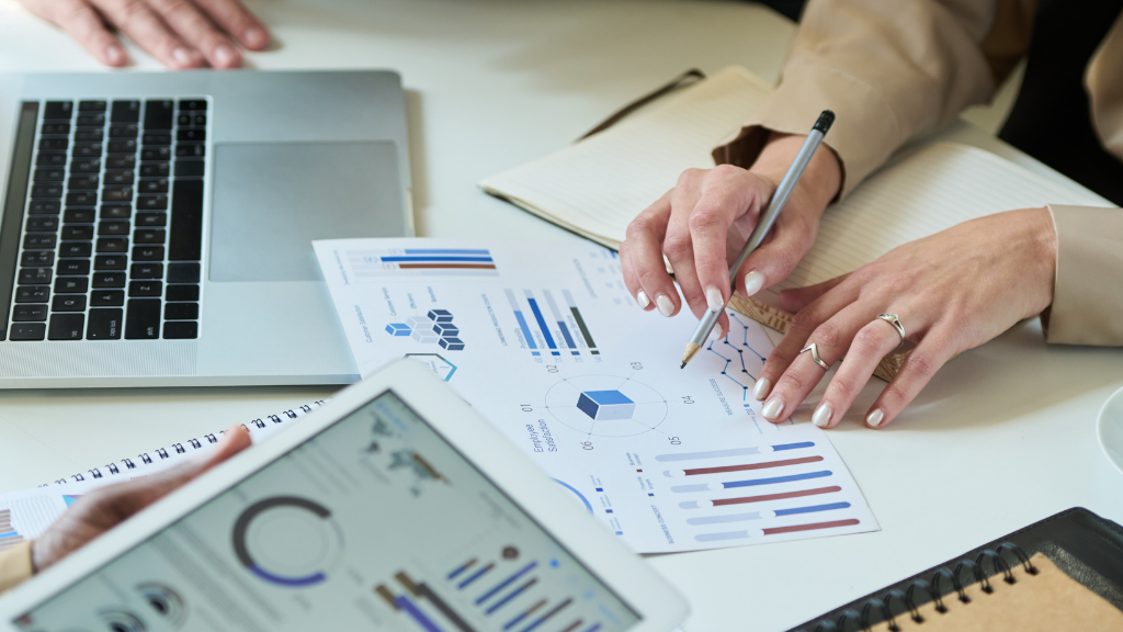 Hands examining financial charts and graphs on paper and a tablet next to a laptop.