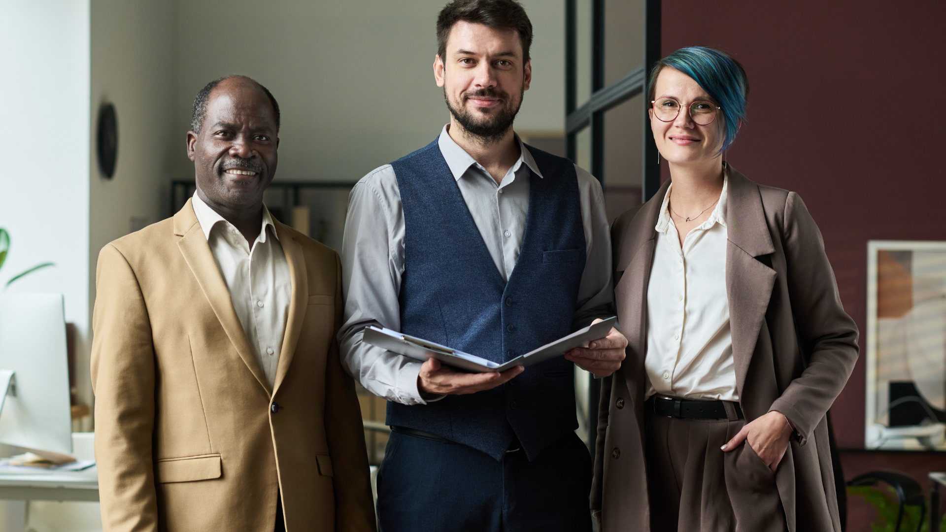 Three people in business attire standing indoors, smiling.