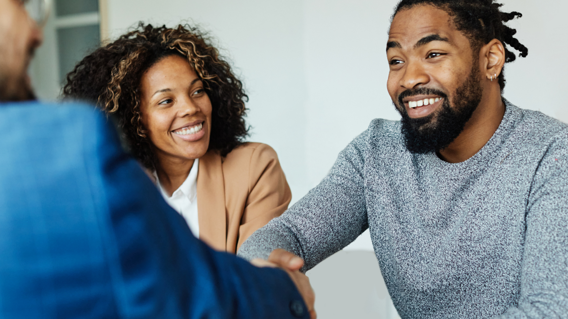 Three people in a meeting: two smiling, one shaking hands. Bright, neutral setting.