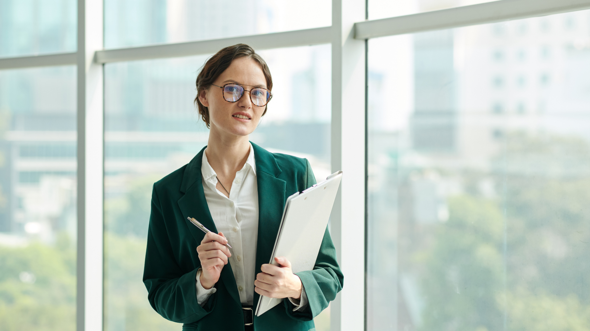 Woman in glasses, green blazer, holding a file and pen, standing near a large window.