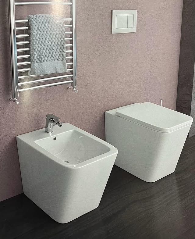In un bagno moderno, un bidet bianco e un WC sospeso accanto a uno scaldasalviette cromato su una parete color malva.
