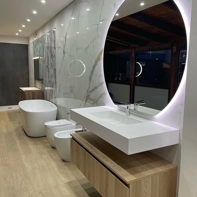 Bagno moderno con specchio rotondo illuminato, mobile lavabo in legno a parete, vasca da bagno bianca e rubinetteria bianca coordinata.