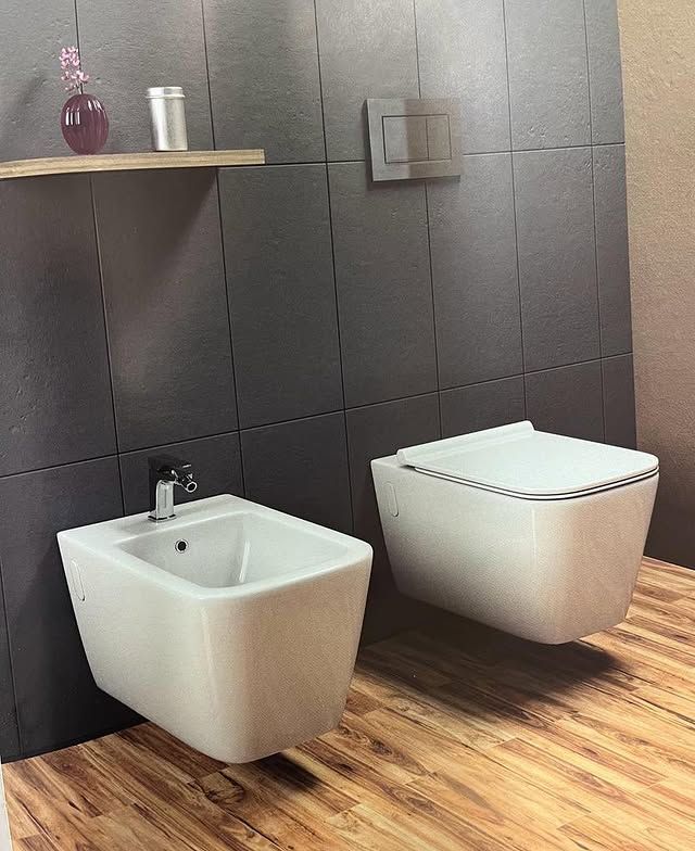 Bidet e WC bianchi a parete, incastonati in una parete piastrellata di scuro sopra un pavimento effetto legno, con mensola in legno sospesa.