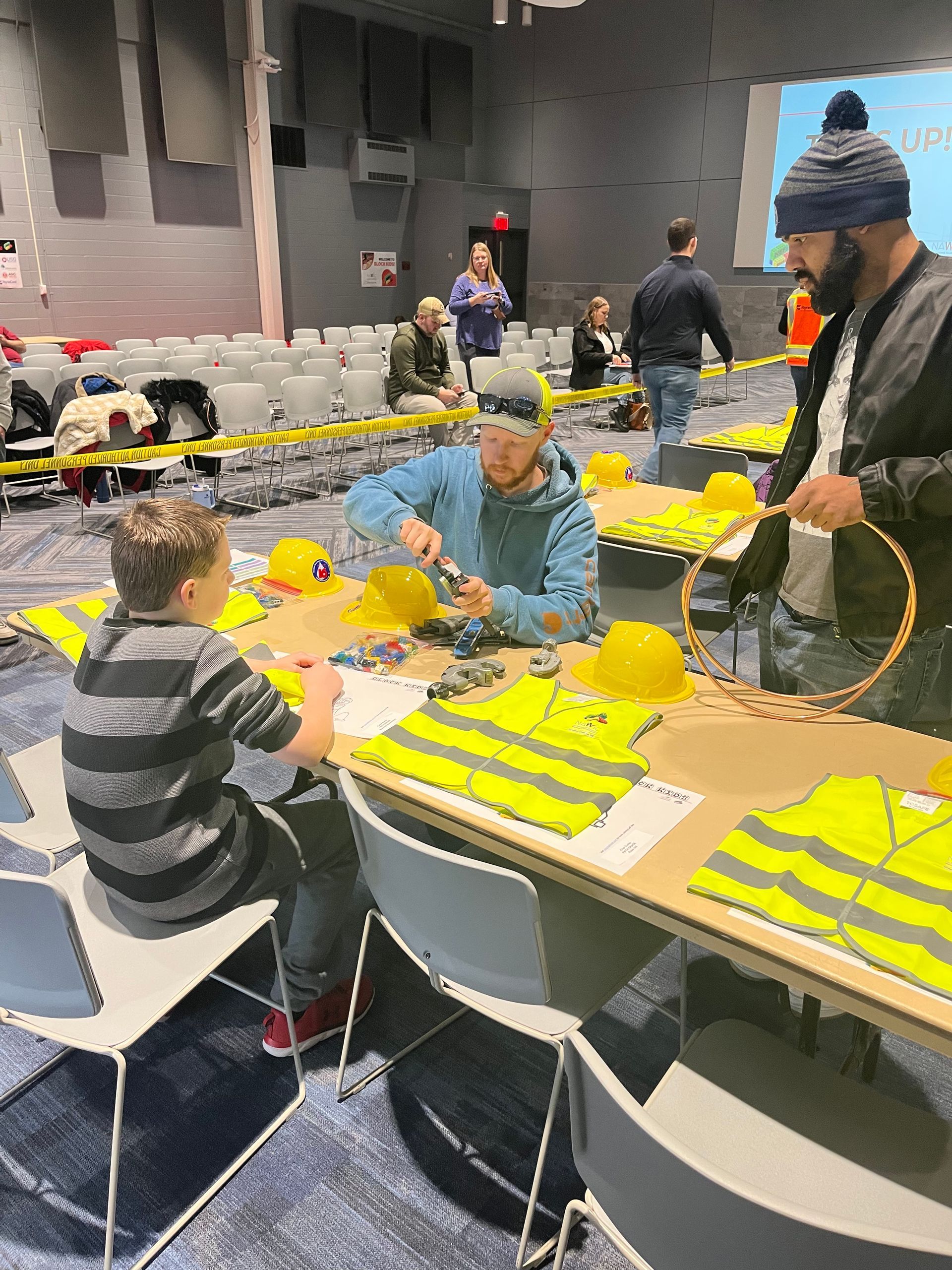 A group of people are sitting at tables in a room making hard hats.