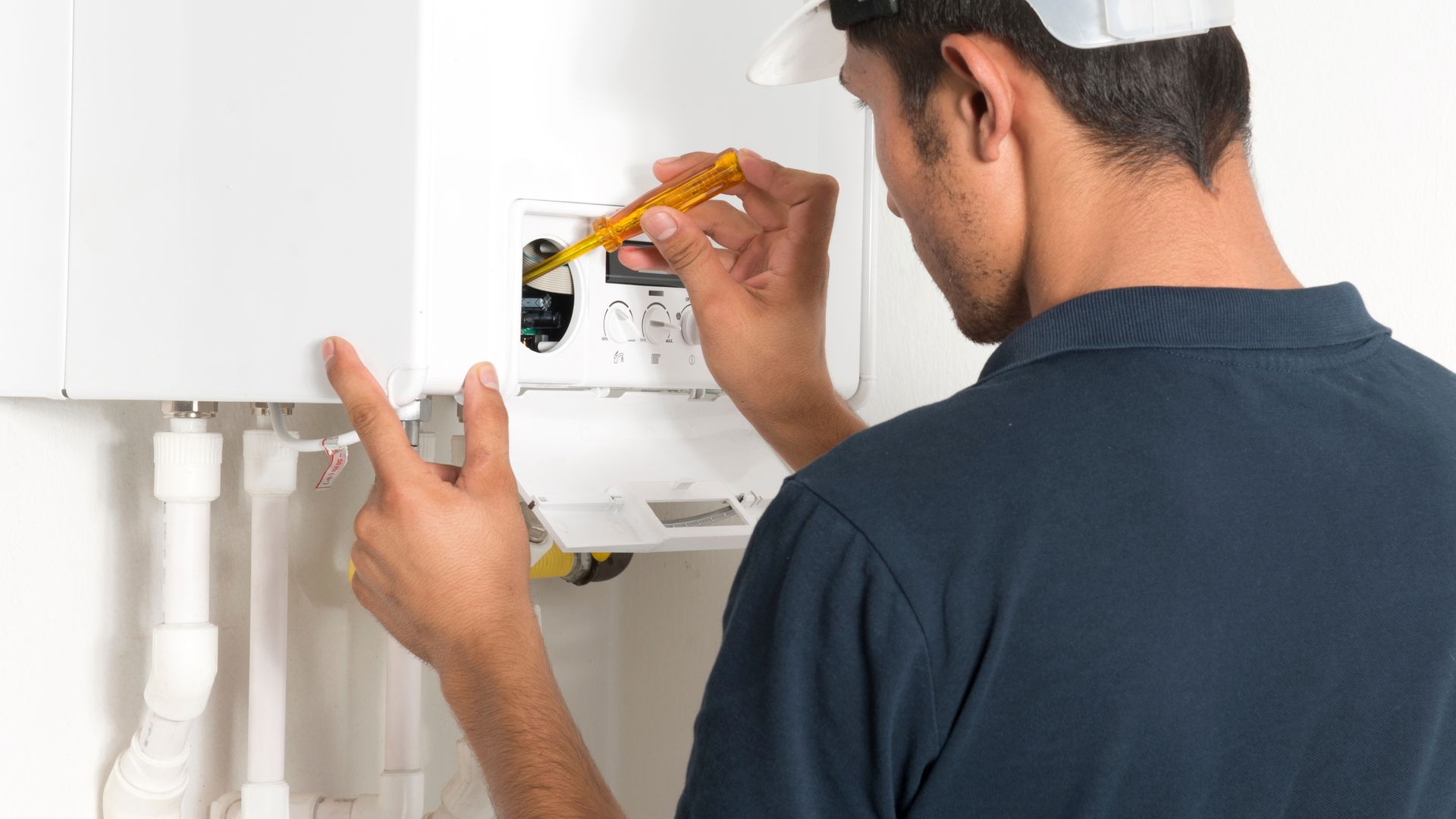 Plumber installing a white wall-mounted boiler with a yellow screwdriver in a bright utility room