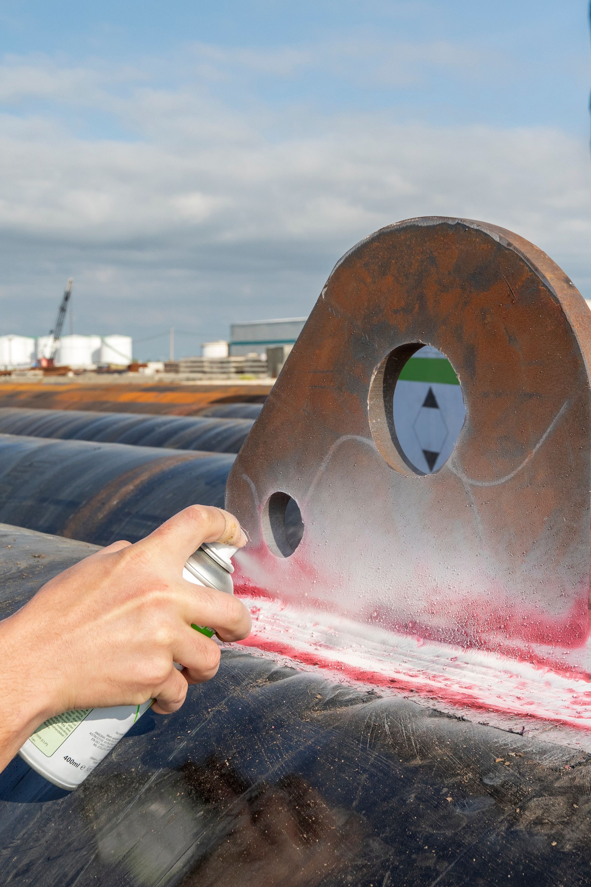 Hand spraying red liquid on a metal component. Steel pipes in the background. Industrial setting.