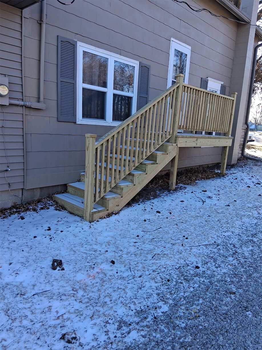 Snow-dusted backyard stairs and wooden railing leading to a house door, beside a window with shutters