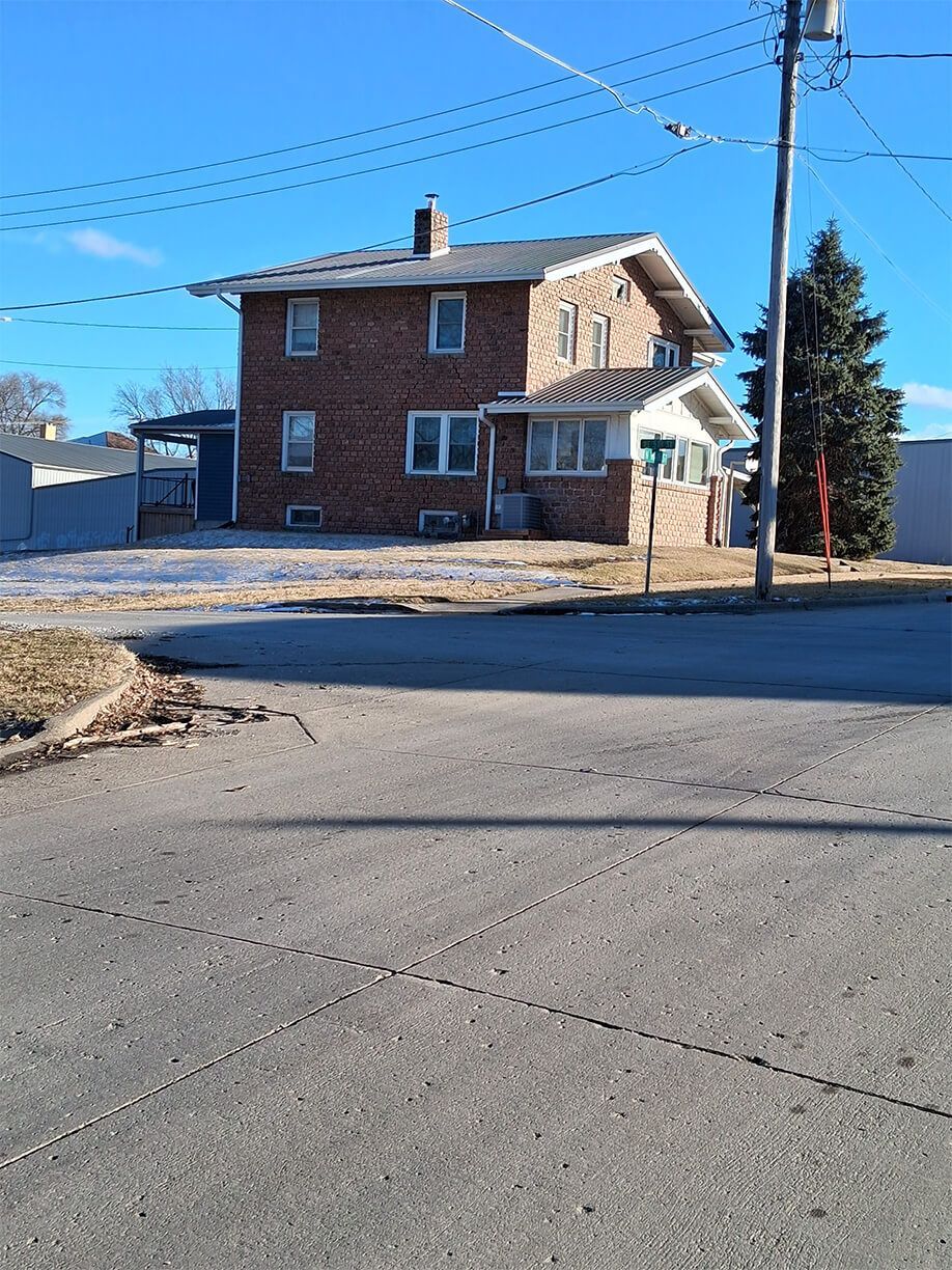 Brick two-story house on a corner lot beside a road, with bare trees and power lines under a blue sky.