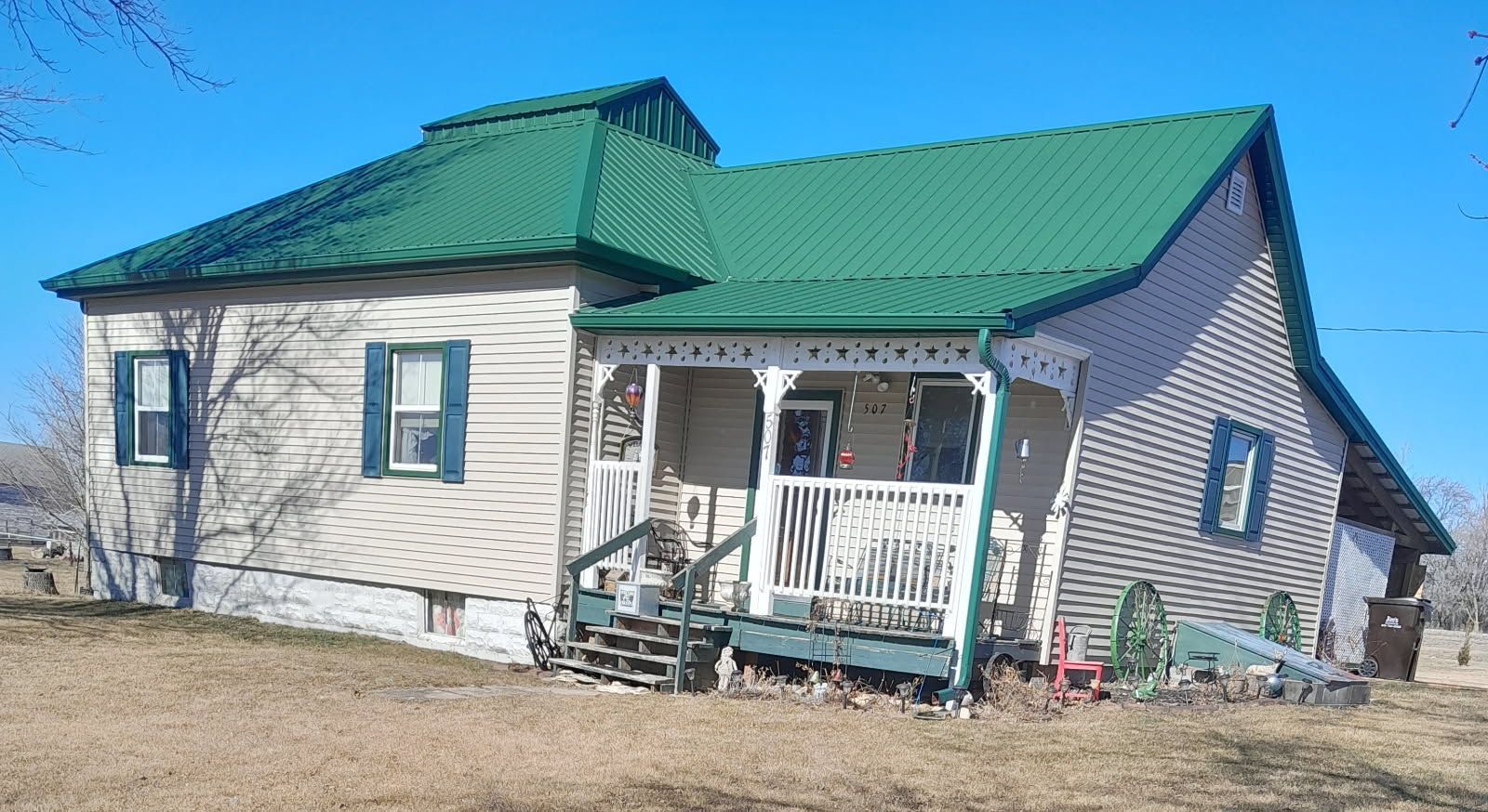 Small house with green metal roof and porch on a bare lot under a clear blue sky.