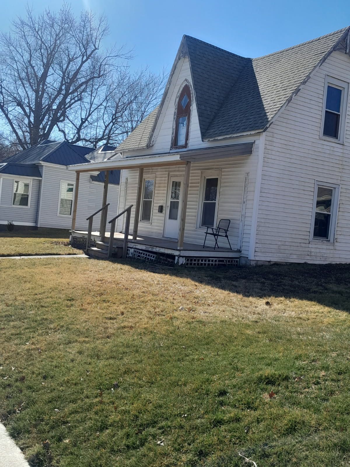 White house with a covered porch and steep roof, sitting on a grassy yard under a clear blue sky.