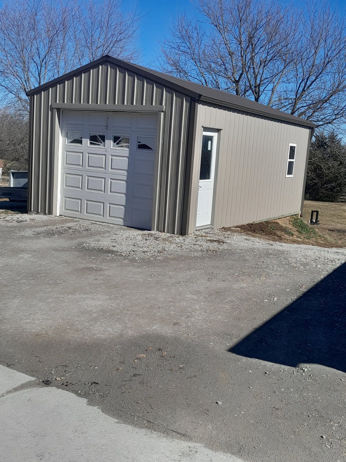 Small beige metal garage with a white roll-up door on a gravel lot under a clear blue sky