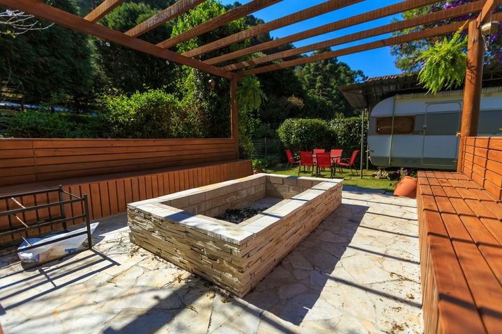 Backyard patio with a stone fire pit, wooden pergola, and red chairs in bright sunlight