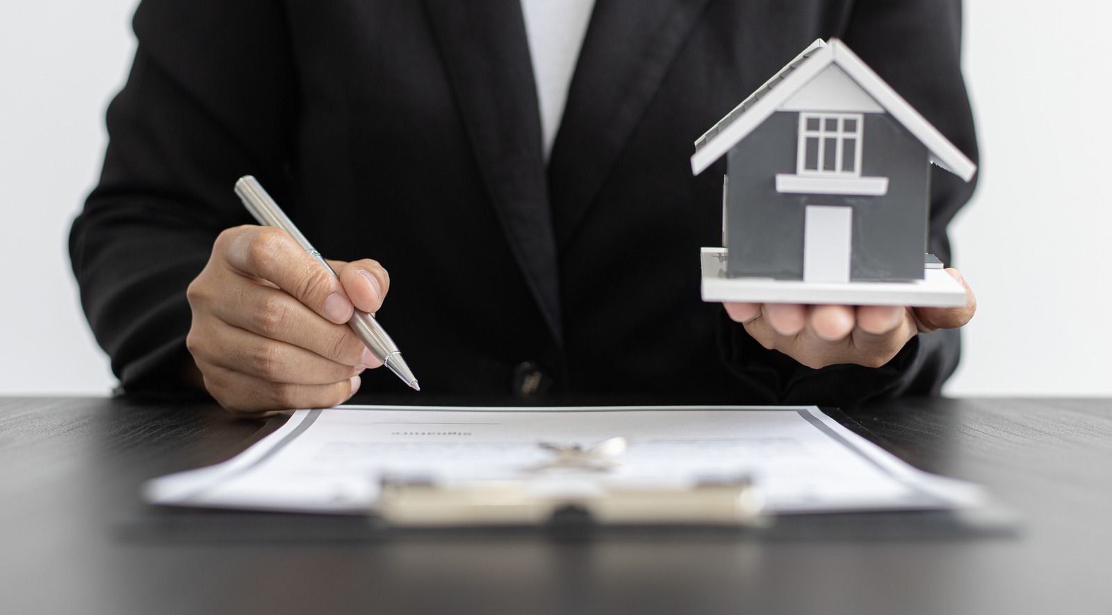 Person in a black suit signs a document beside a small house model