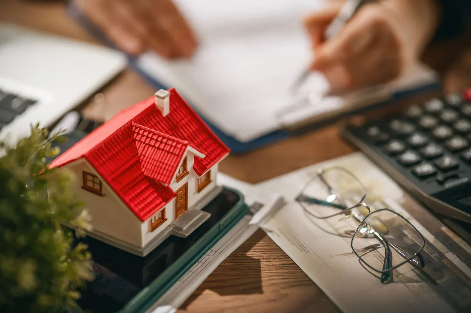 Miniature house with a red roof on a desk beside eyeglasses, calculator, and hands working in the background