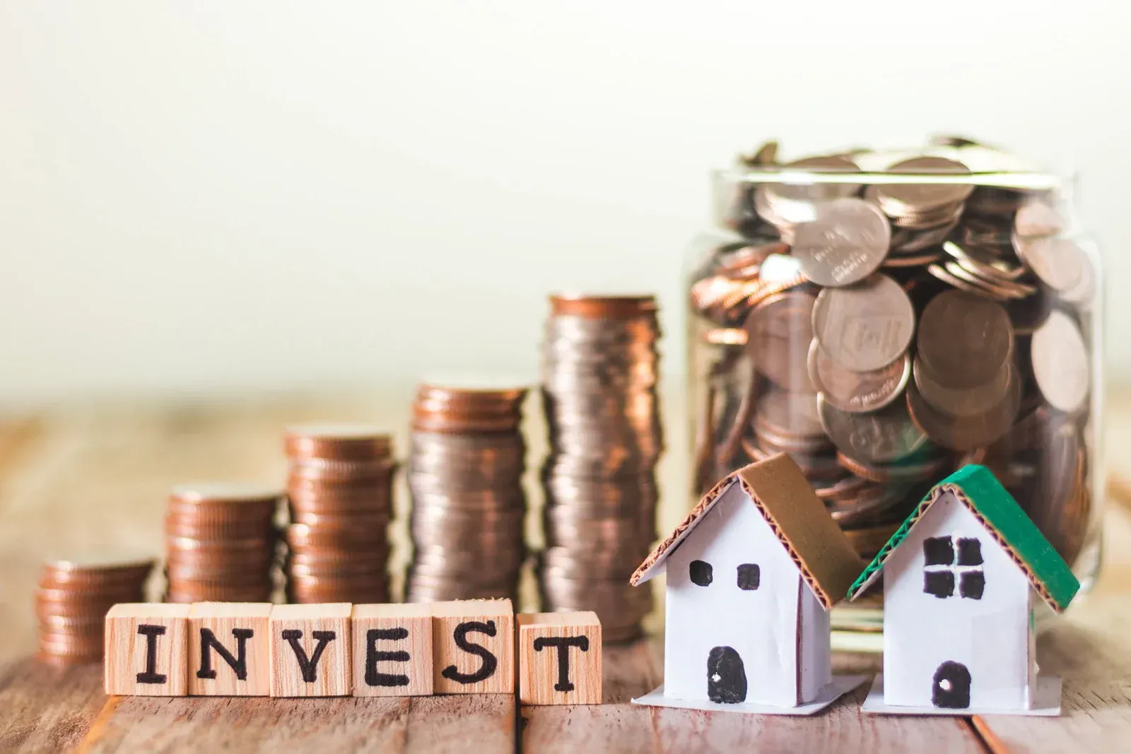 Stacks of coins, house models, and wooden blocks spelling “INVEST” on a table