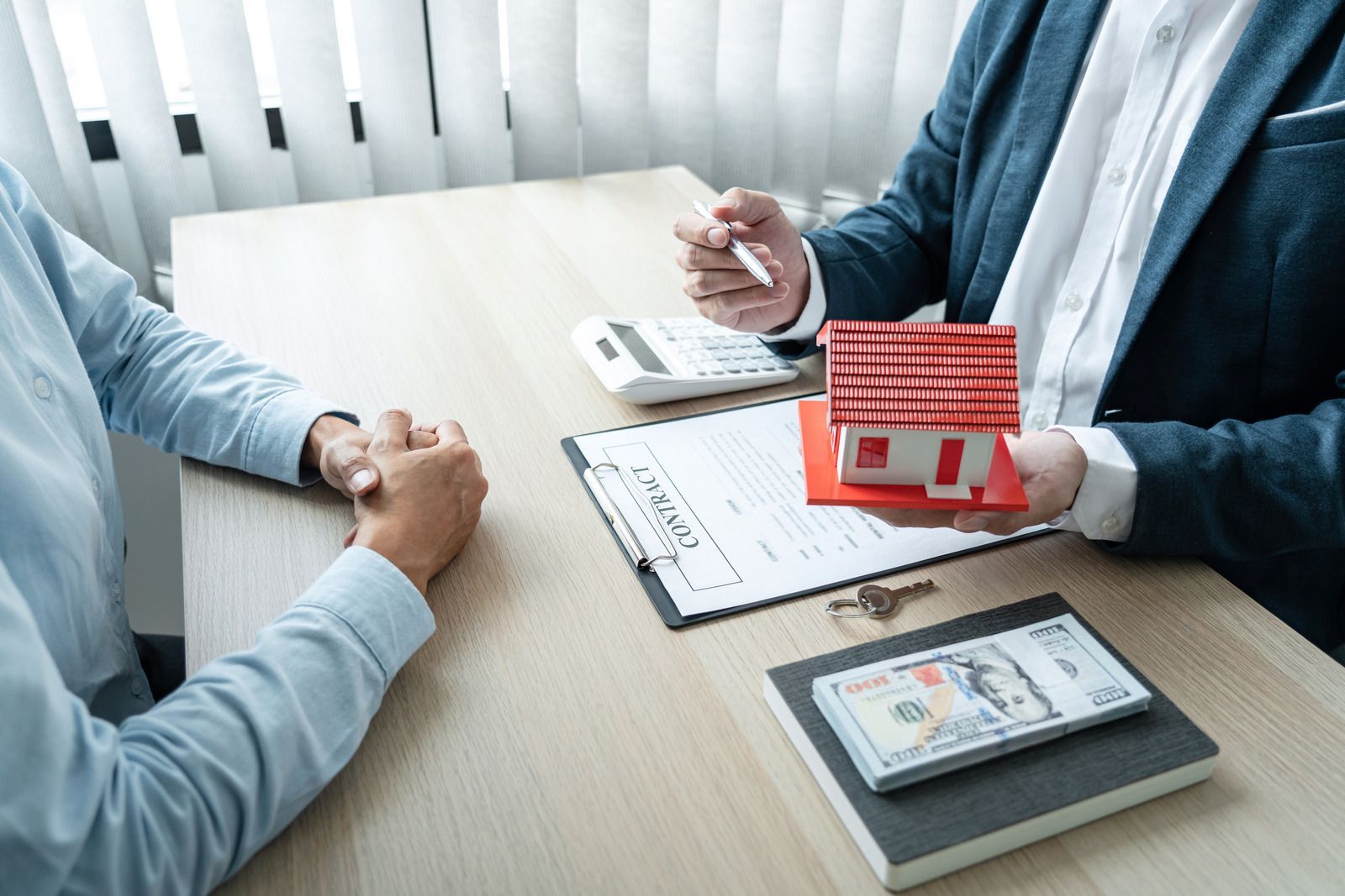 Two people meeting at a desk with paperwork, calculator, and a red model house beside a dollar bill booklet.
