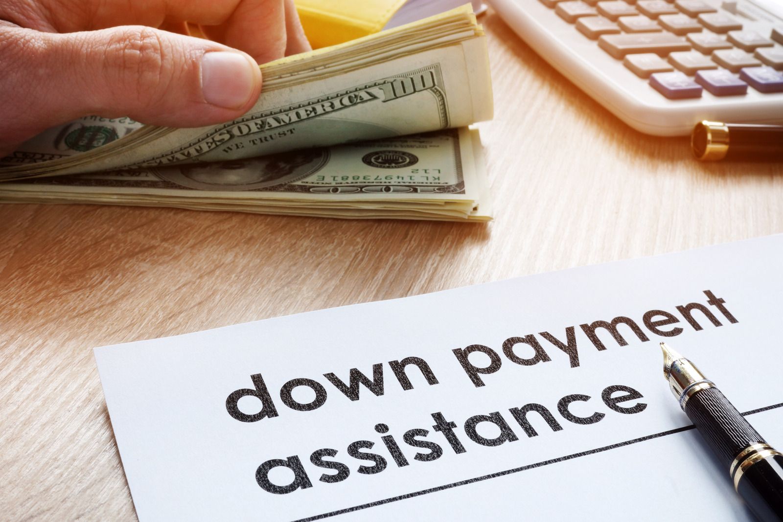Money held above a paper labeled “down payment assistance” beside a pen and calculator