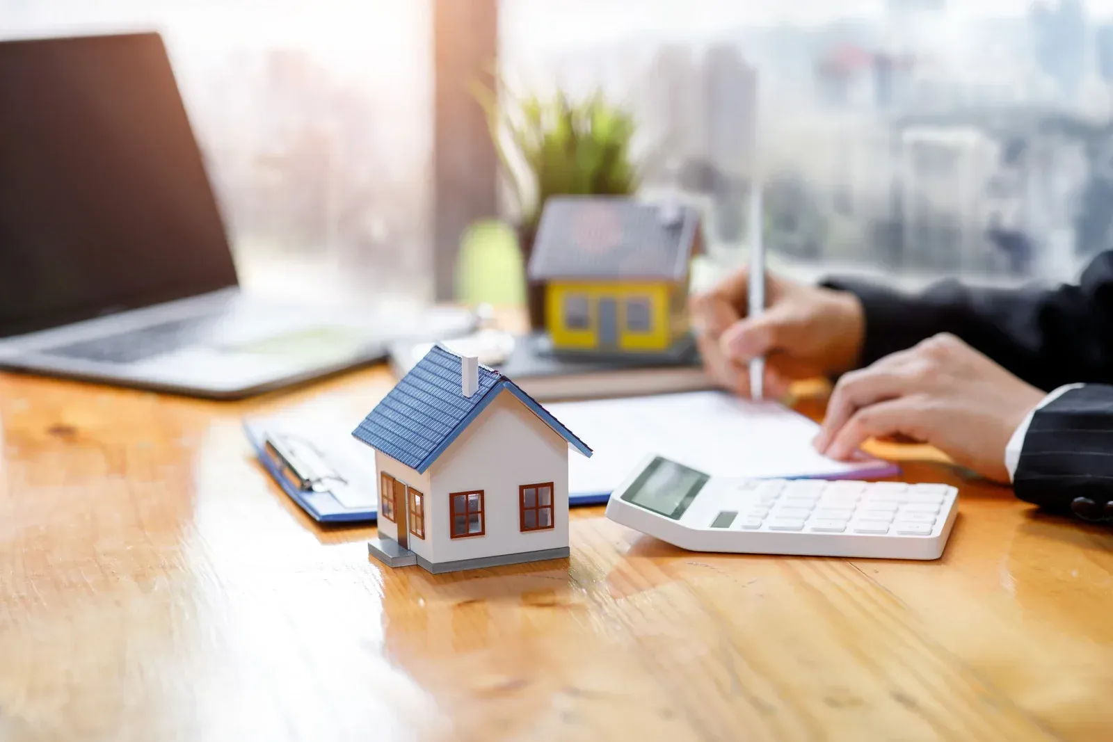 Miniature house on a desk with calculator, laptop, and a person writing in the background.