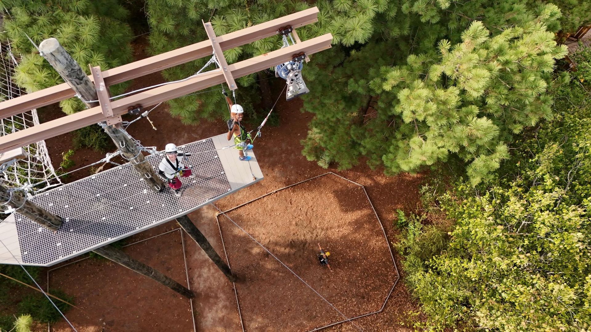 People on a ropes course platform in a wooded area, viewed from above.