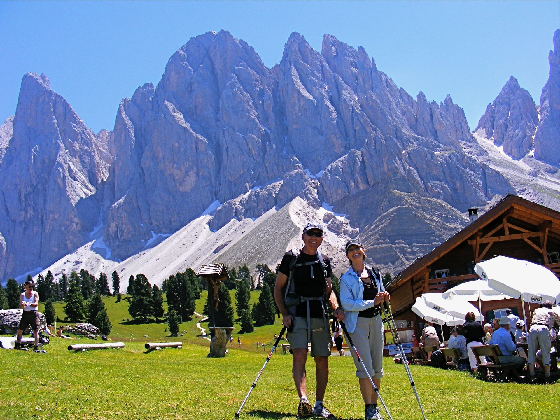 Two hikers with trekking poles pose in front of the Dolomites. Green grass, blue sky, and a wooden hut are visible.