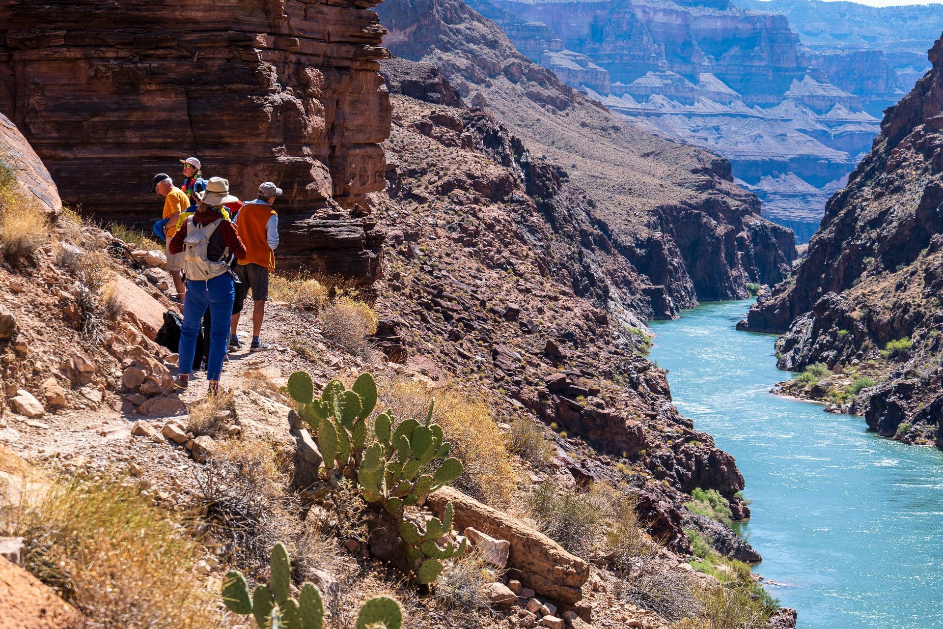 Hikers on a rocky canyon trail overlooking a blue river between steep cliffs.