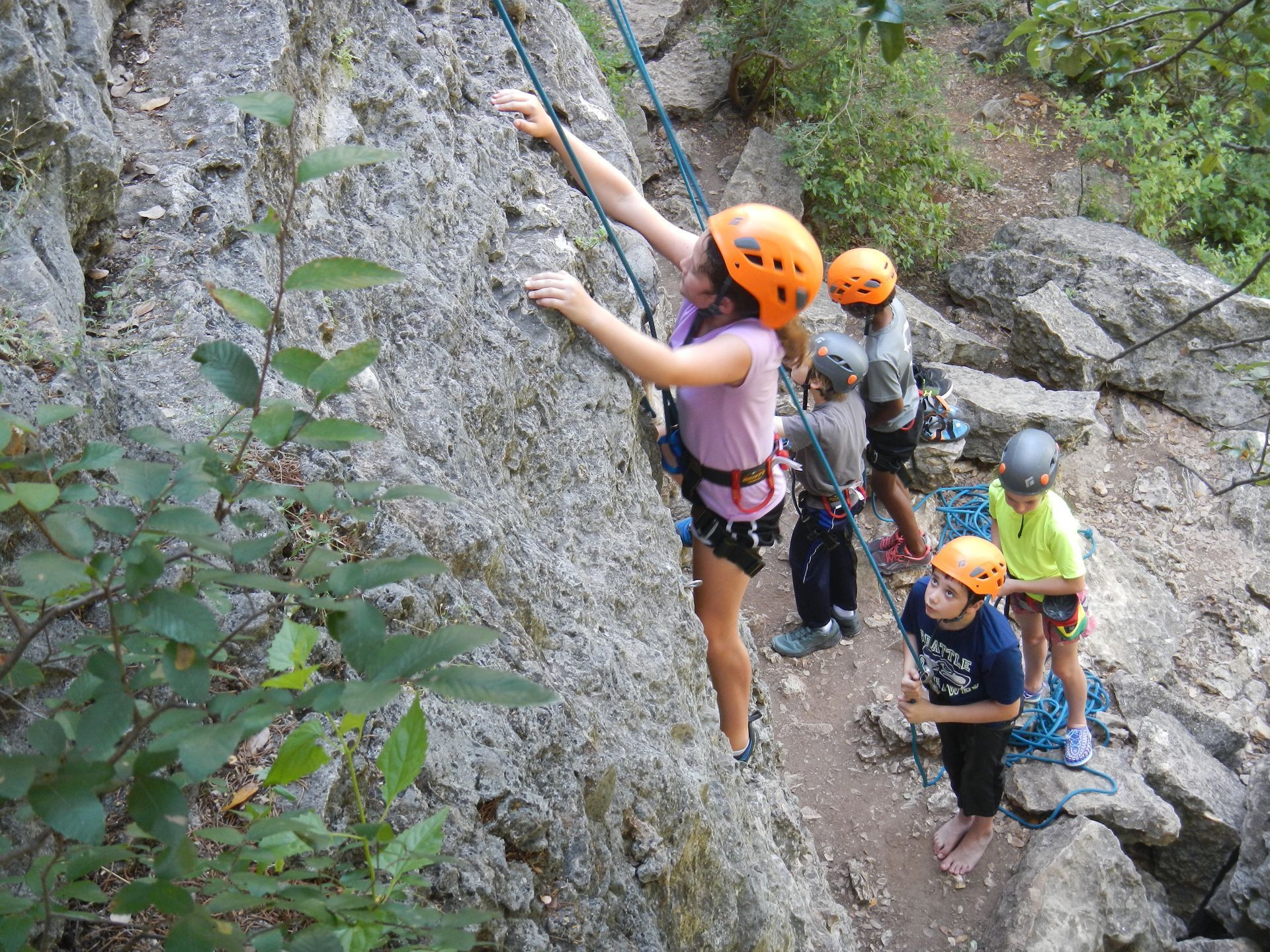 Climbers in orange helmets scaling a steep rocky cliff on a narrow trail through greenery