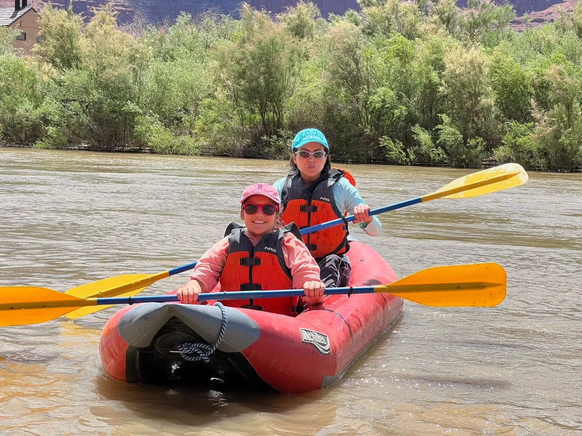 Two people in red life jackets paddling a red kayak on a river with yellow oars