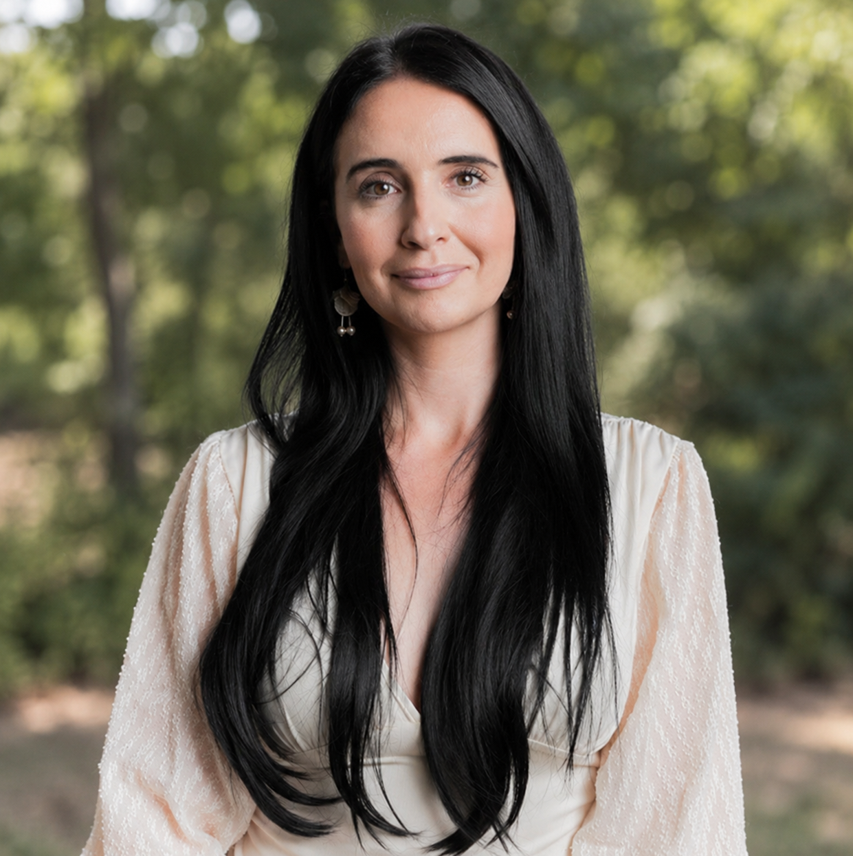 Portrait of a woman with long black hair in a light blouse, standing outdoors with blurred greenery behind her