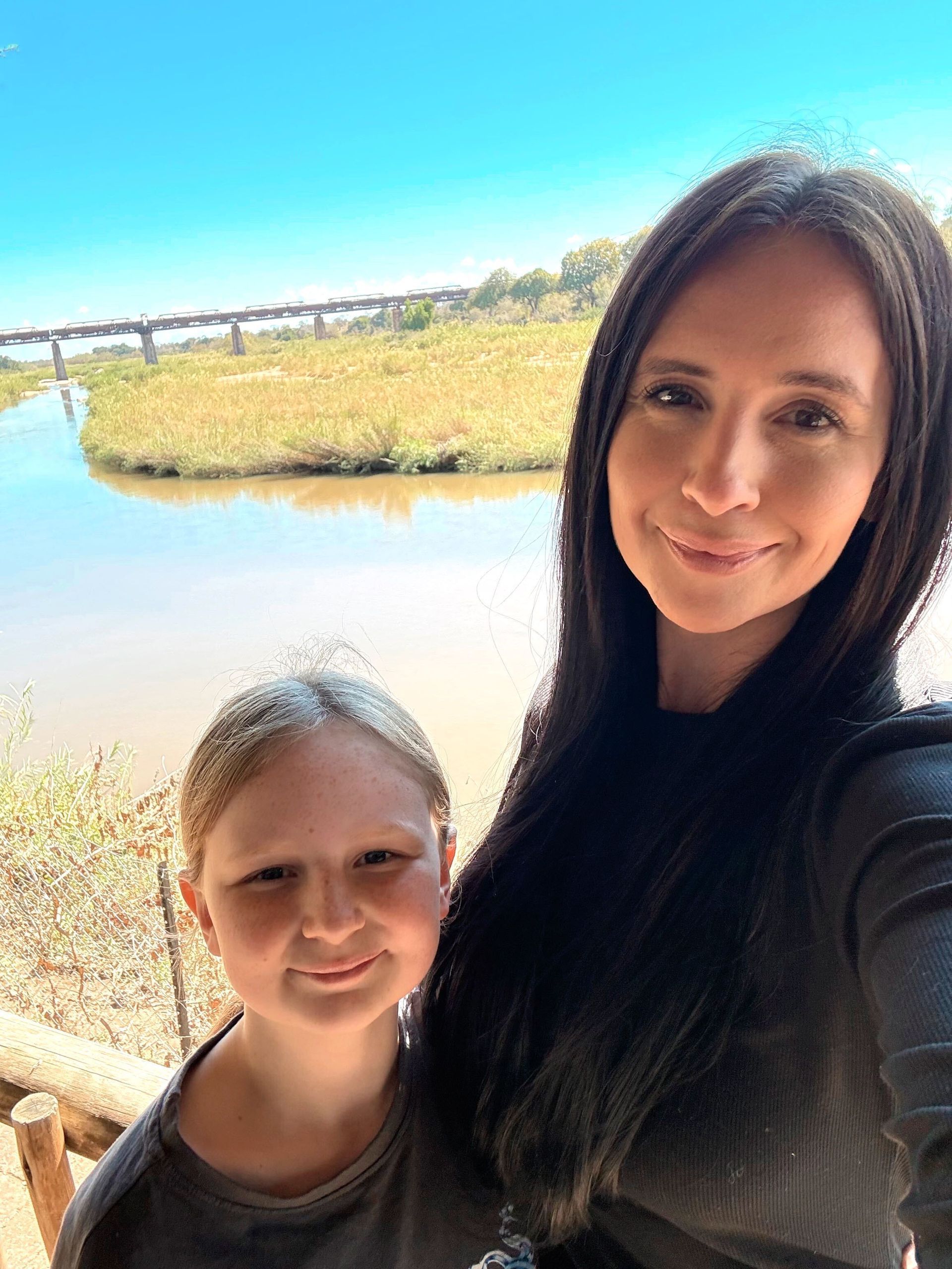 Two people smiling by a bright lakeside with water, reeds, and blue sky in the background