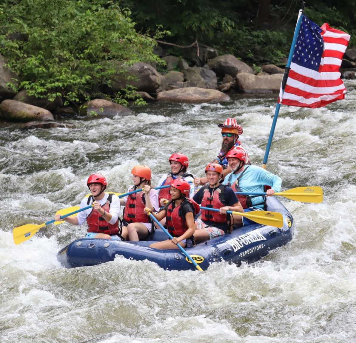 Rafting group on whitewater rapids with yellow paddles and an American flag, surrounded by rocky riverbanks