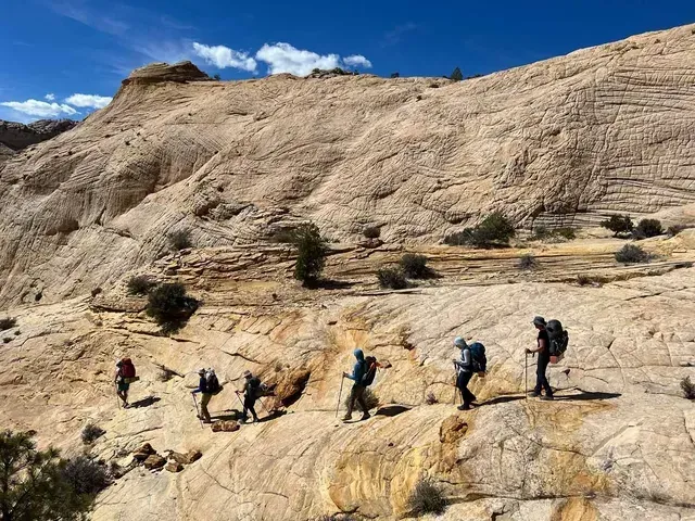 A line of six backpackers hiking across a sloping, light-colored rock formation under a bright blue sky.