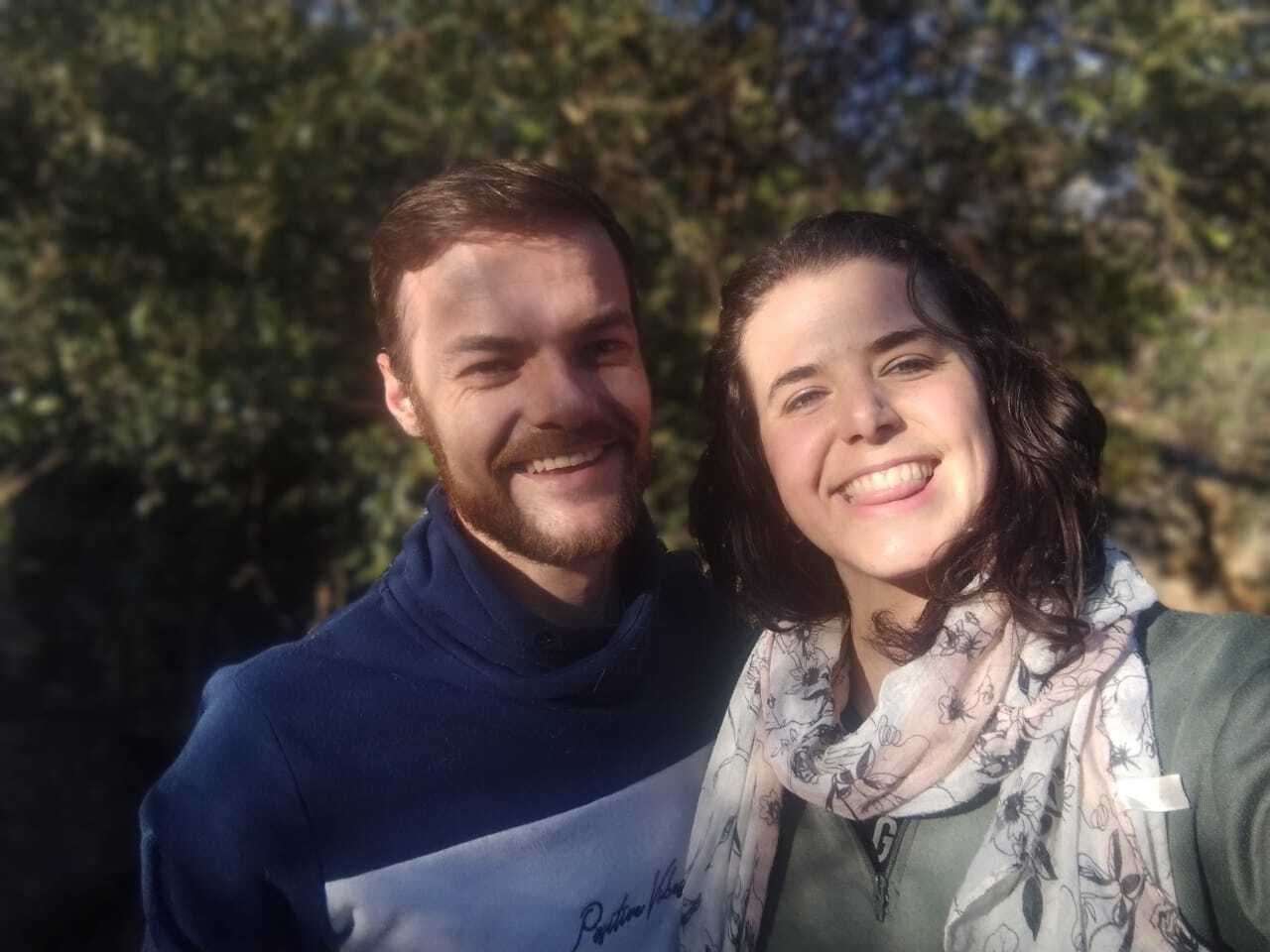 Two smiling people outdoors in sunlight, standing close together with trees in the background.