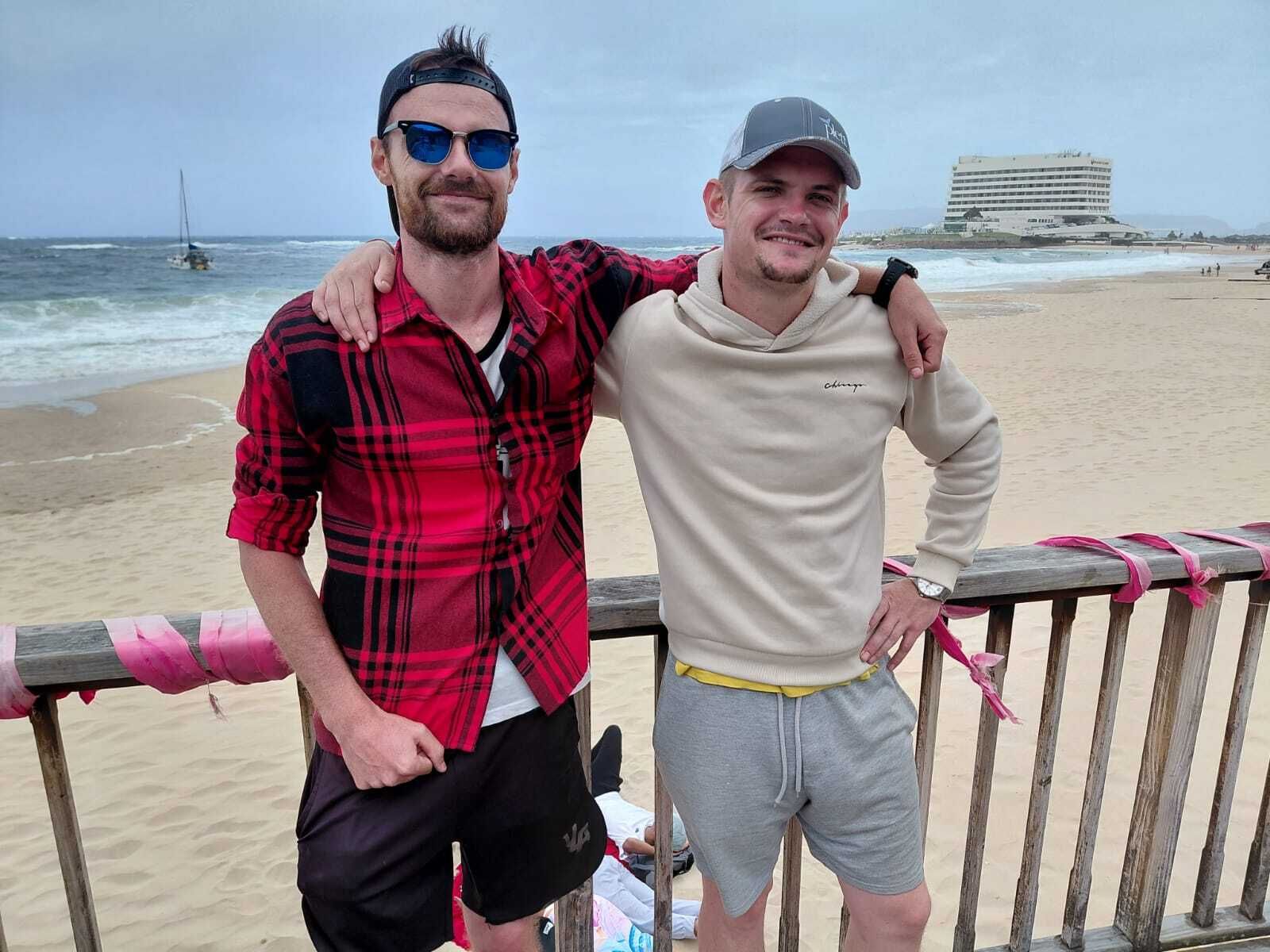 Two friends posing by a beach railing, smiling in casual clothes with the ocean behind them