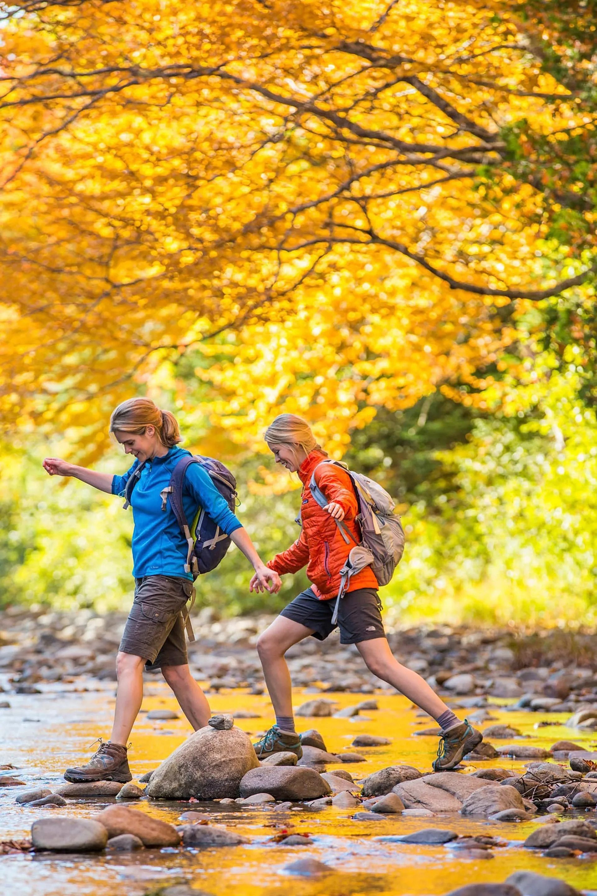 Two hikers in colorful jackets crossing a shallow stream under bright autumn trees