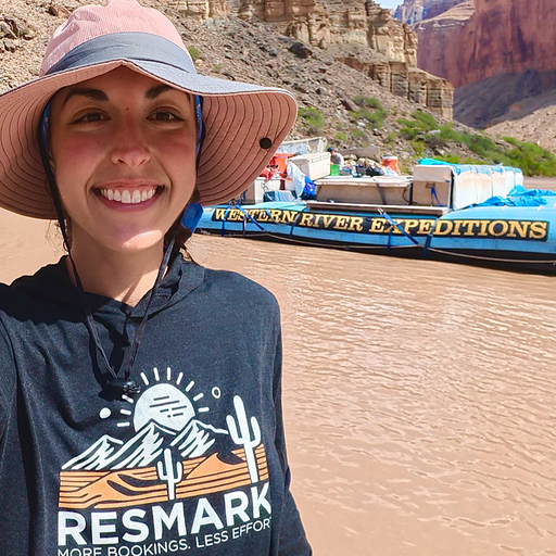 Smiling person in a sun hat stands by the Colorado River near a whitewater raft in a canyon