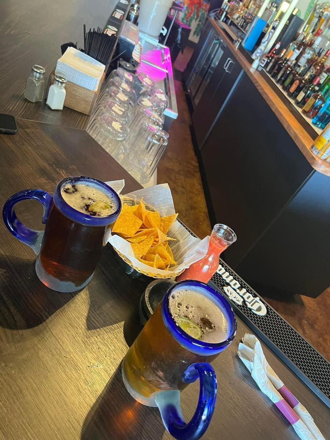 Two large beer mugs with blue handles, chips, and a cocktail on a bar.