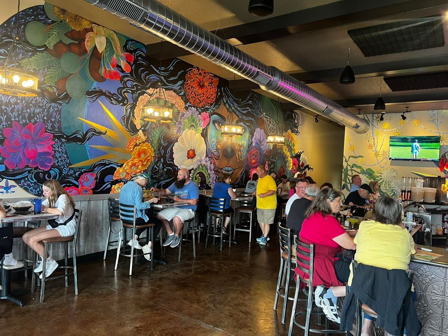 A crowded restaurant with a floral mural; people eat and drink at tables and the bar.