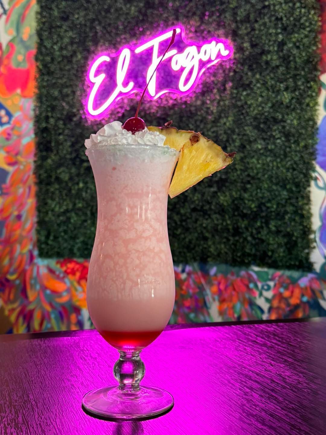 Pink cocktail in a tall glass, topped with whipped cream and cherry, garnished with pineapple slice; neon sign