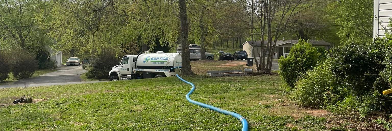 A white water tanker truck parked on a grassy area, connected to a hose. Lush green trees surround it.