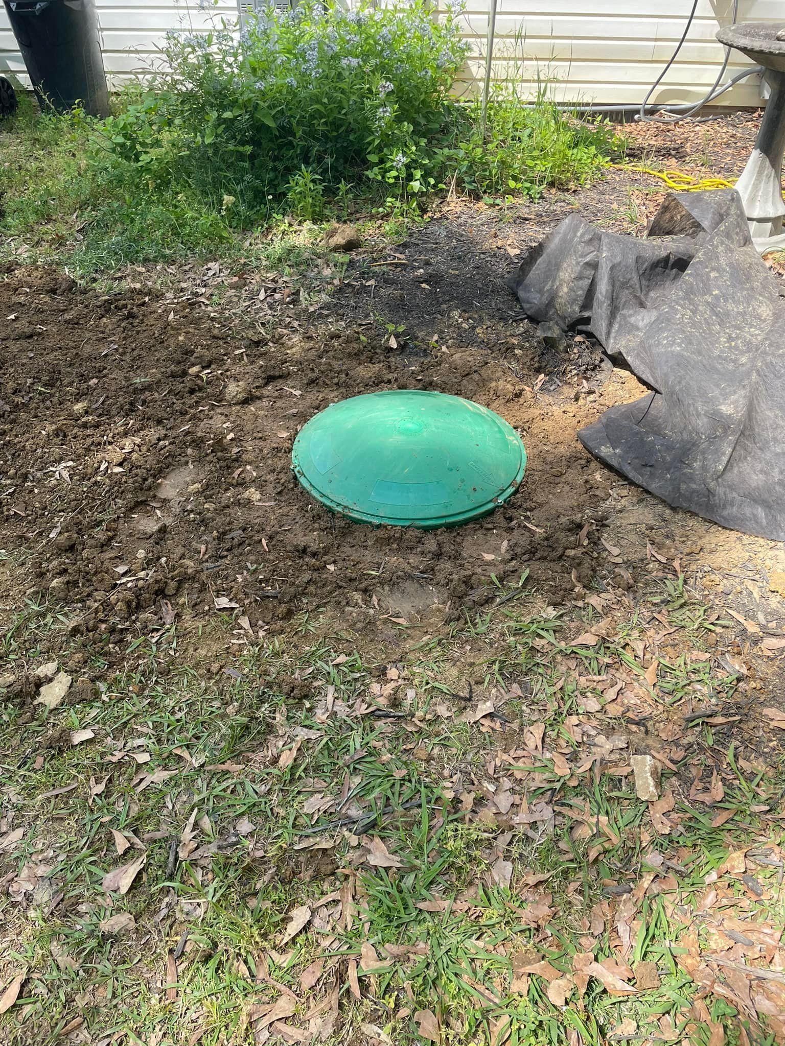 Green septic tank lid in a muddy yard, near overgrown bushes.