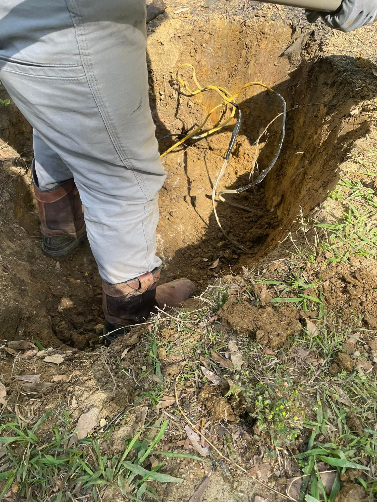 Person in light pants and boots in a dirt trench, holding wires. Sunny outdoor setting.